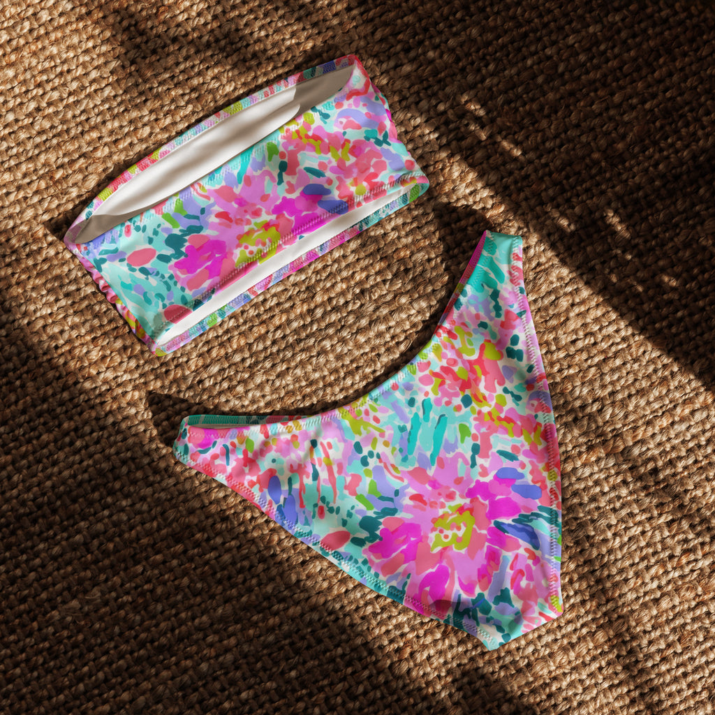 Abstract preppy print recycled bandeau bikini top and V-cut bottoms laid flat on a woven surface