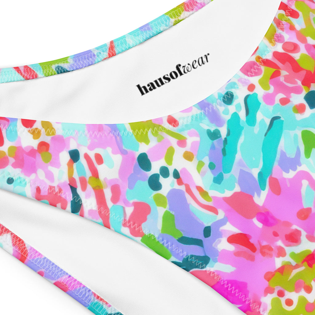 Abstract preppy print recycled bandeau bikini top and V-cut bottoms laid flat on a woven surface